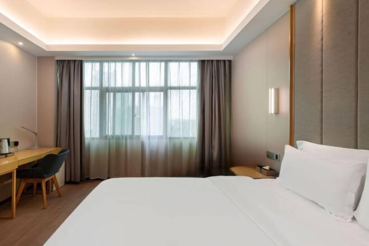 Ein Hotelzimmer mit einem großen Bett, einem Schreibtisch und einem Stuhl. Es gibt ein Fenster mit Vorhängen und einen Nachttisch mit einer Lampe.