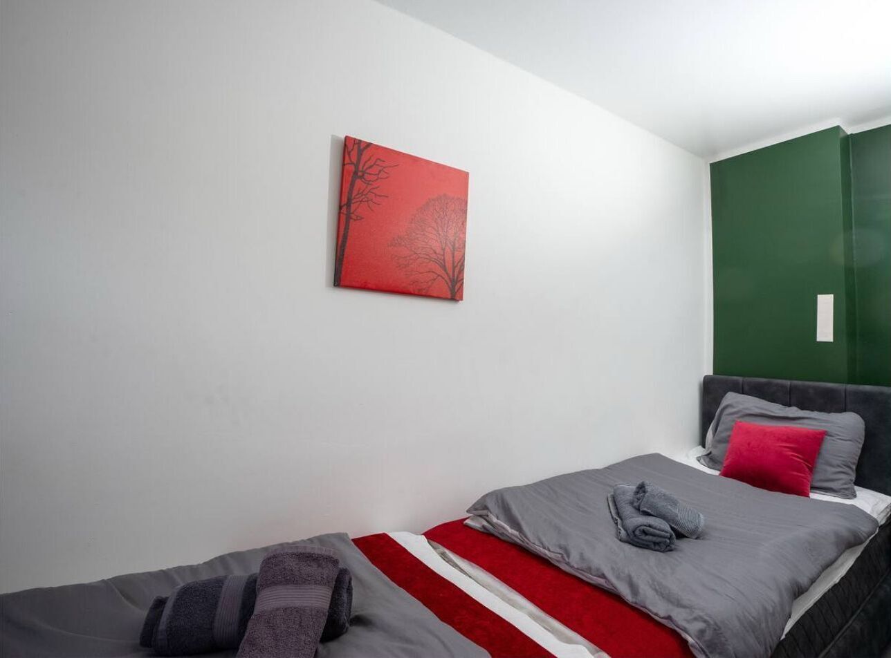 A bedroom with two beds, one with a red and white striped blanket and the other with a grey blanket. There is a red painting on the wall and a green accent wall.