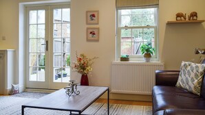 Ein Wohnzimmer mit großem Fenster, Heizkörper, einer Couch, einem Couchtisch und zwei gerahmten Bildern an der Wand.