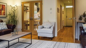 Ein Wohnzimmer mit einer Couch, einem Sessel, einem Couchtisch und einem Essbereich mit Stühlen und einem Tisch. Durch eine offene Tür ist eine Küche zu sehen.