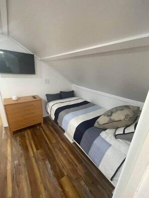 Une petite pièce étroite, avec un lit simple, une commode en bois et un téléviseur à écran plat fixé au mur.