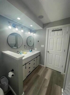 Une salle de bain dotée d’un grand meuble-lavabo, de deux miroirs ronds, d’un éclairage au plafond, d’un porte-serviettes et d’une porte sur laquelle est inscrit « SERVIETTES ».