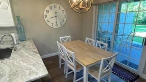 A kitchen with a marble countertop, a wooden dining table with white chairs, a wall clock, and a chandelier.