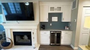 A compact living space with a fireplace, a television mounted above it, and a kitchen area with white cabinets and a stainless steel refrigerator.
