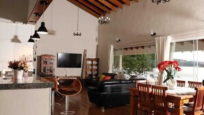 Ett rymligt vardagsrum med en svart lädersoffa, ett matbord och stolar i trä. En platt-TV är monterad på väggen, och det finns bokhyllor fyllda med böcker.