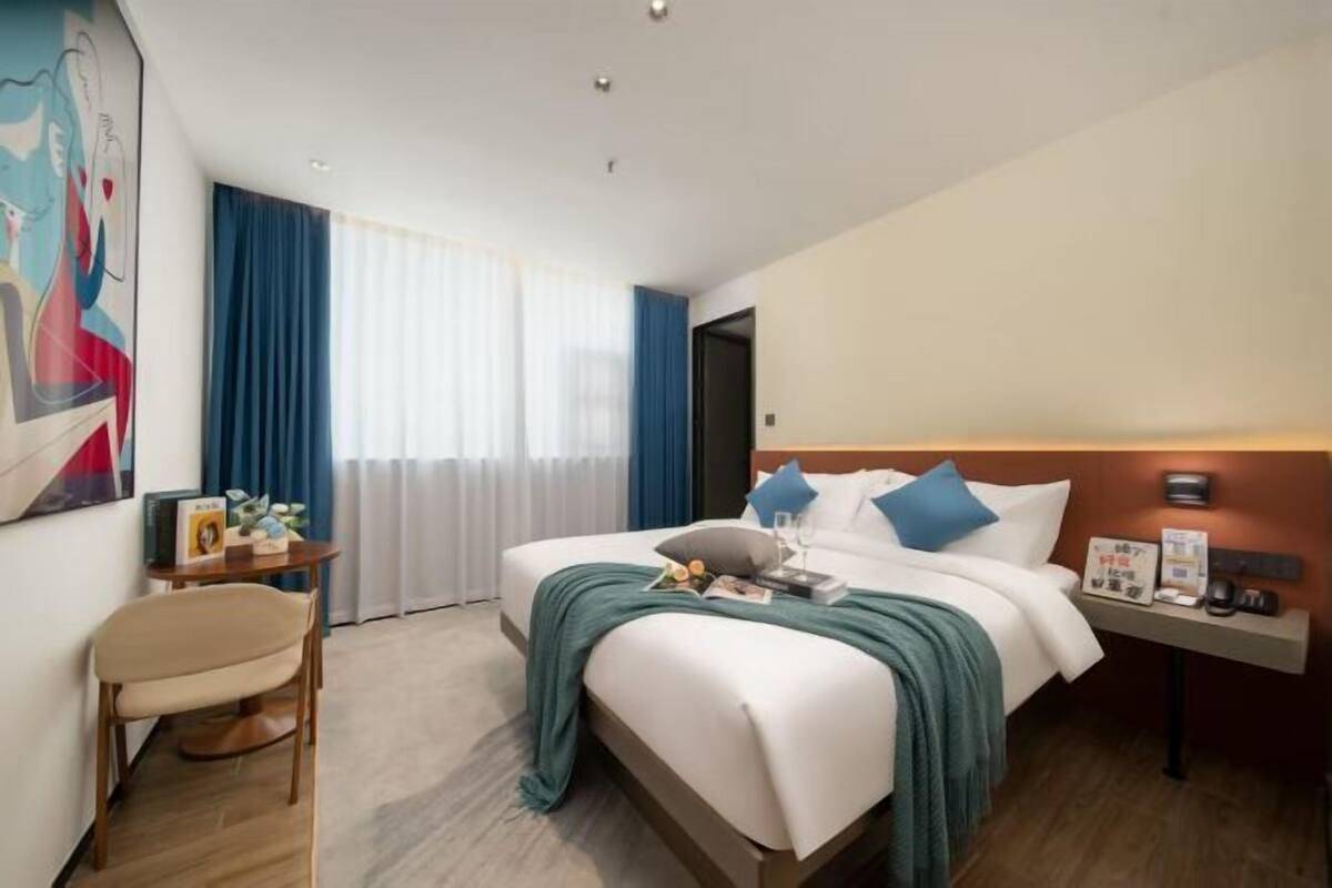 A hotel room with a bed, a bedside table, a chair, a window with curtains, and a painting on the wall.
