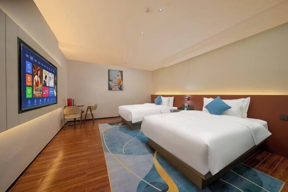 A hotel room with a large digital display, two beds, a chair, and a painting on the wall.