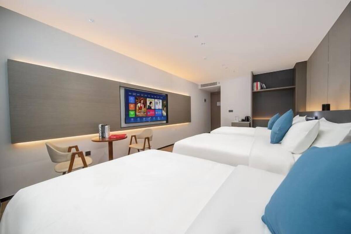 A modern hotel room with two beds, a TV mounted on the wall, a small table with a chair, and a bookshelf.