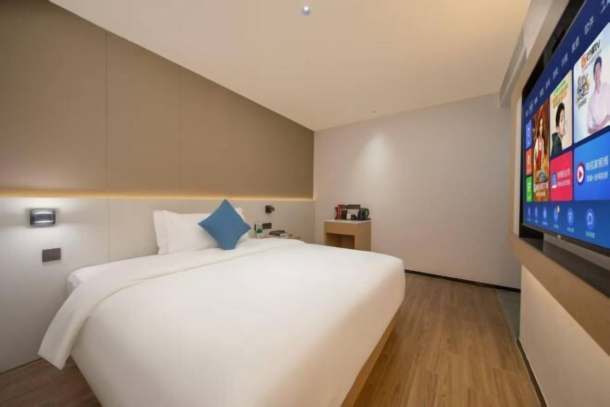 A modern hotel room with a large bed, a bedside table, a TV mounted on the wall, and a wooden floor.