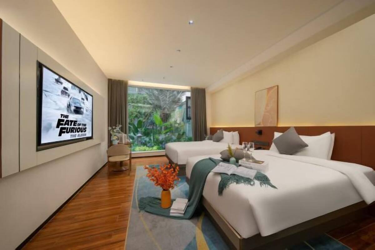 A hotel room with two beds, a large window with a view of greenery, a poster on the wall, and a small table with a vase of flowers.