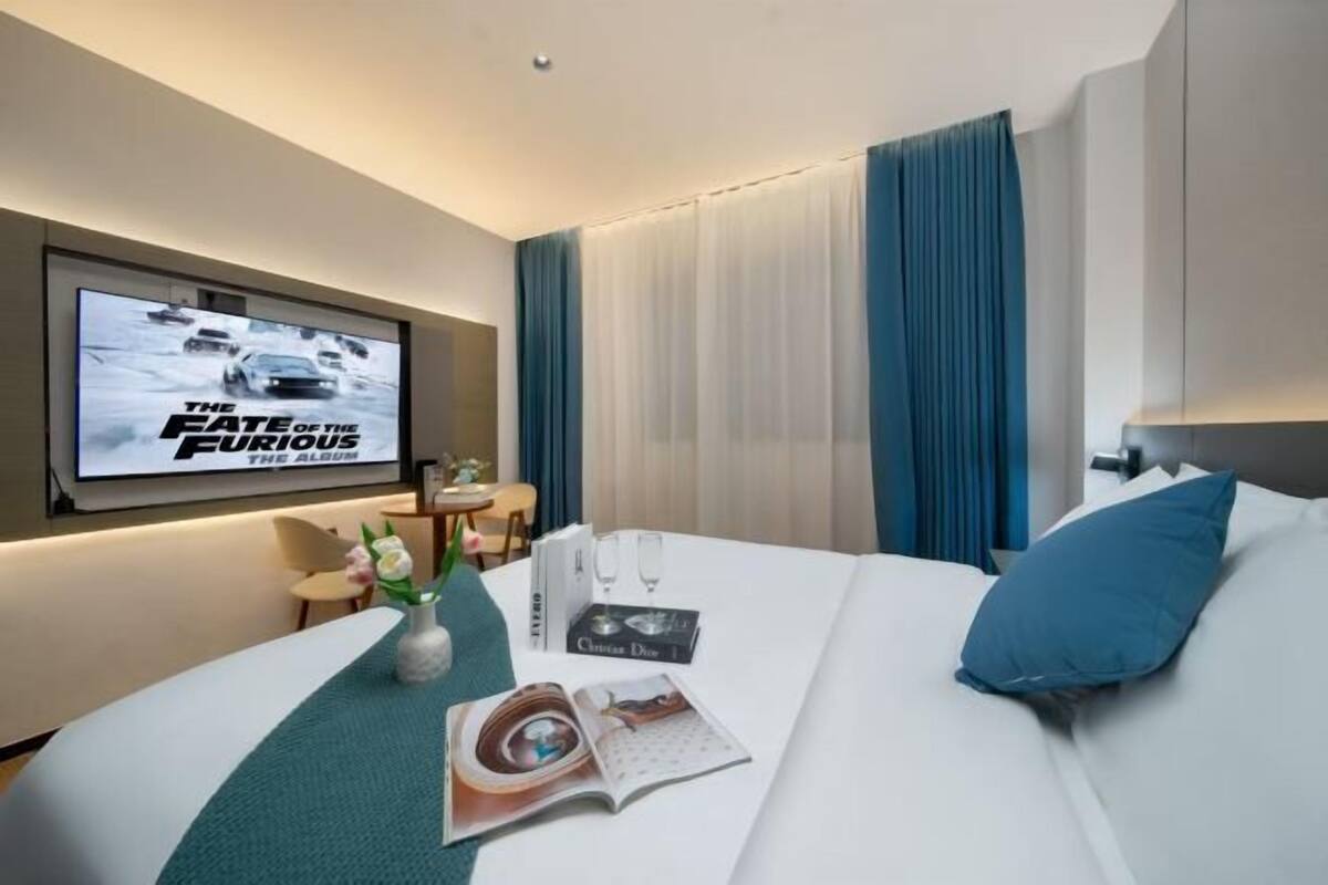 A hotel room with a bed, a TV displaying 'The Fate of the Furious,' a book, and a vase with flowers.