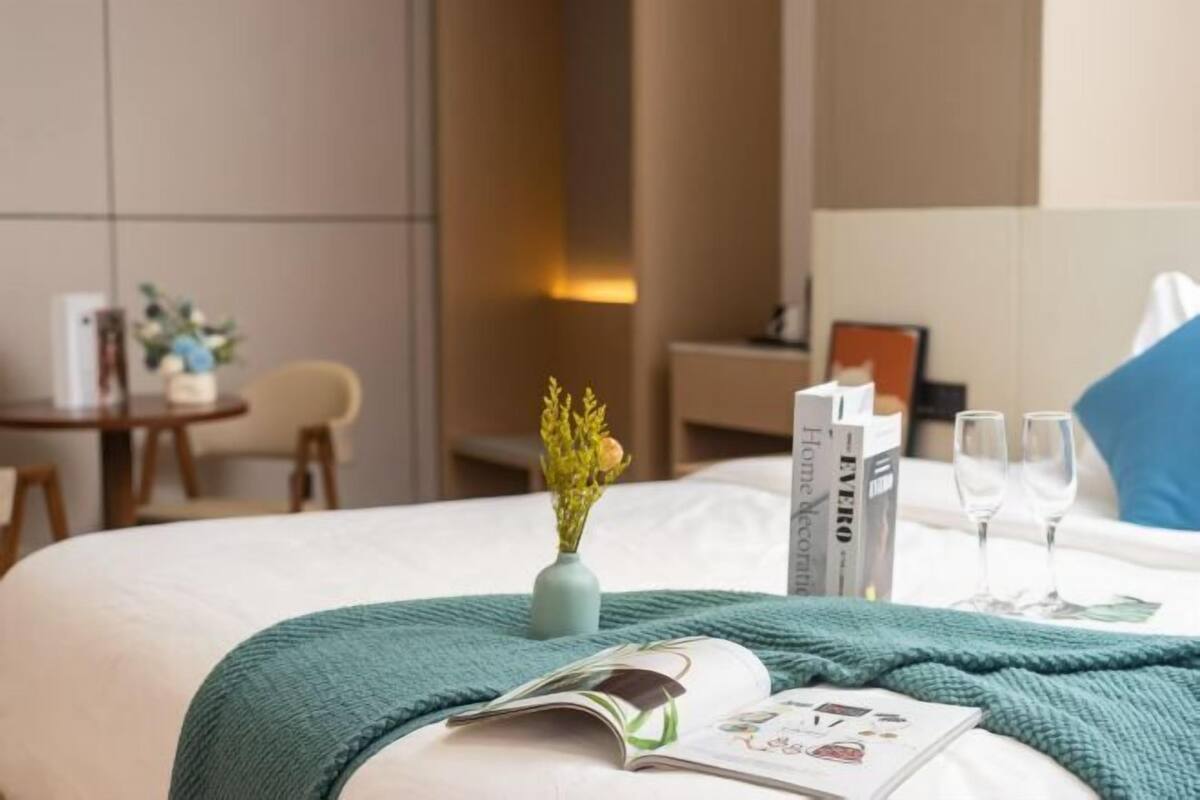 A neatly made bed with a green blanket, a vase with flowers, a magazine, and two glasses on the side.