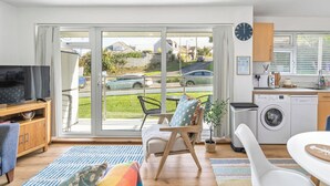 A modern living room with a television, sofa, and a dining area with a table. There is a sliding glass door leading to a balcony with a view of the street and cars. - 2 bedroom accommodation in Newquay (Newquay)