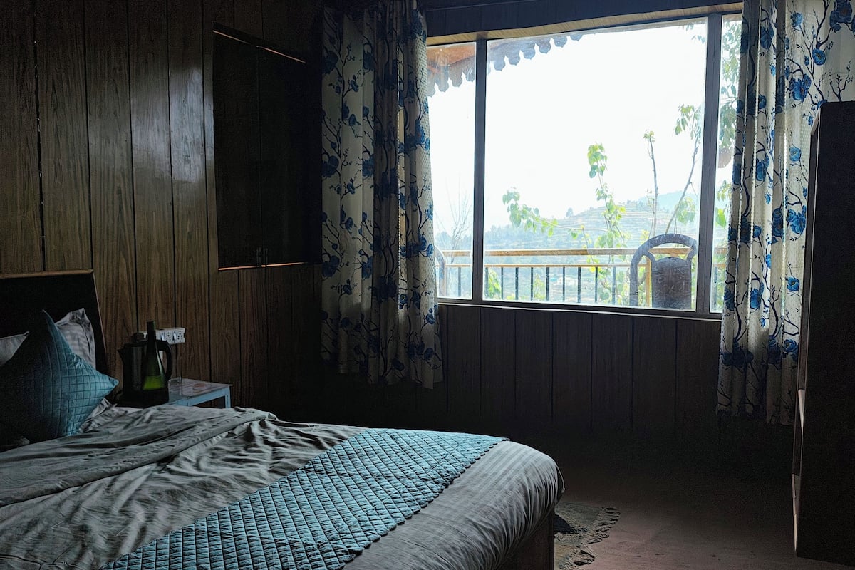 A bedroom with a bed, a nightstand, a window with curtains, and a view of the outdoors.
