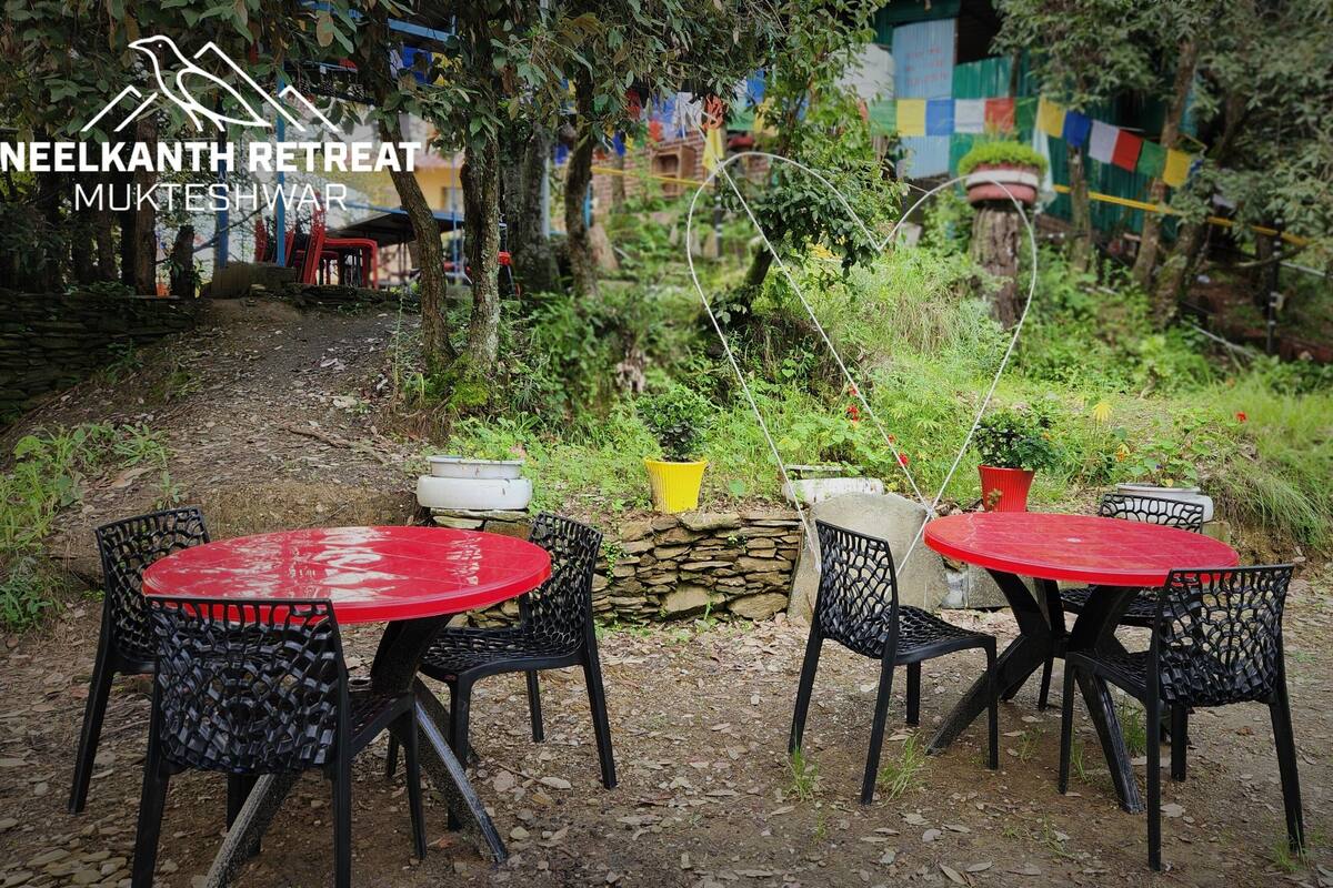 An outdoor seating area with red tables and black chairs, surrounded by greenery and stone walls.
