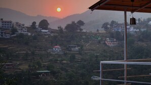 Udsigt fra en balkon på Neelkanth Retreat i Mukteshwar, med solnedgang over et bakket landskab med terrasseret jordbrugsmark og bygninger.