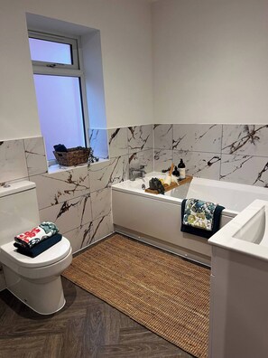A bathroom with a toilet, a bathtub, a window, and a woven mat on the floor. - Long Shoot - Beautiful, spacious and luxurious house. (Nuneaton)