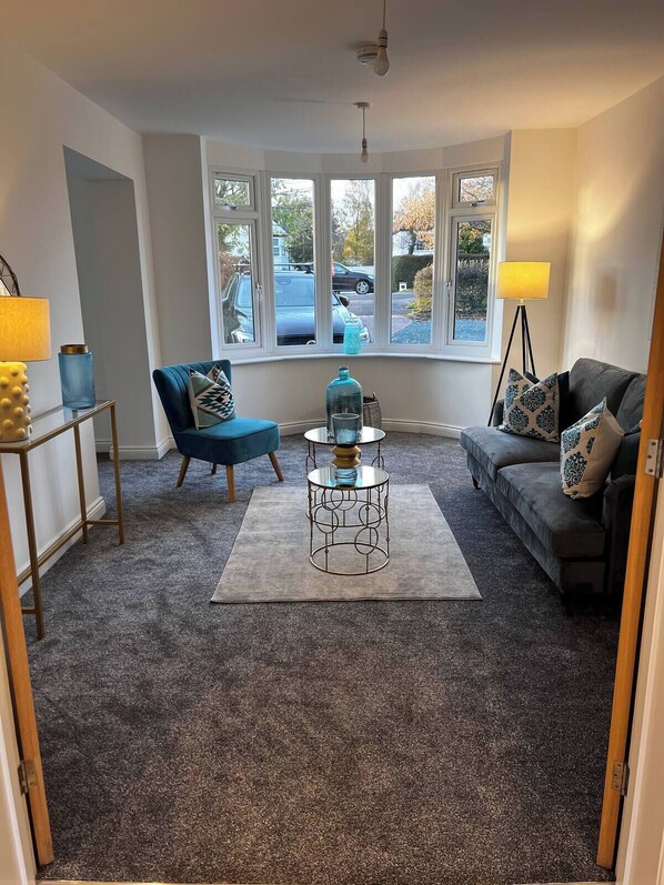 A living room with a blue armchair, a grey sofa, a coffee table, and a view of parked cars outside through large windows. - Long Shoot - Beautiful, spacious and luxurious house. (Nuneaton)