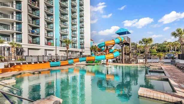 Une piscine d’hôtel avec un toboggan, un immeuble à plusieurs étages doté de balcons et des palmiers.
