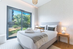 A modern bedroom with a large bed, a bedside table with a lamp, a tray with breakfast items, and a view of greenery through the sliding glass door. - Babi by the Sea by Atlantic Holiday (Funchal)