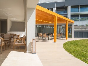 An outdoor seating area with wooden tables and chairs, a covered section with a yellow roof, and a modern building in the background. - Babi by the Sea by Atlantic Holiday (Funchal)