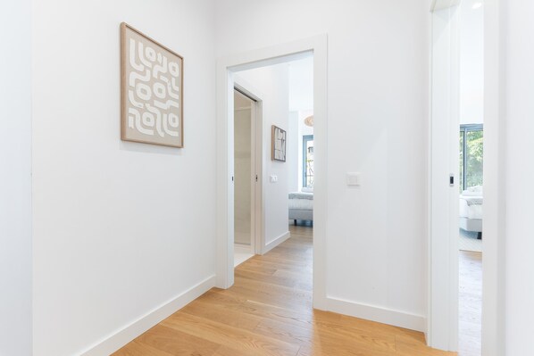 A hallway with wooden flooring and white walls, leading to a bedroom with a bed and a window.