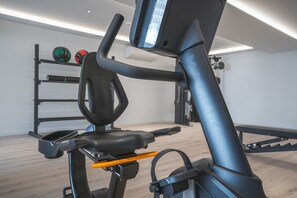 En stillesiddende cykel med digital skærm, et vægmonteret stativ med træningsbolde og en bænk.