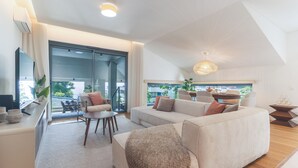 A modern living room with a sofa, coffee table, and a TV on a stand. There is a sliding glass door leading to an outdoor area with a view of greenery. - Babi by the Sea by Atlantic Holiday (Funchal)