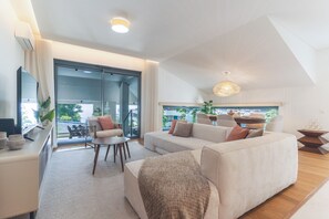 En moderne stue med sofa, sofabord og fjernsyn på et stativ. Der er en skydedør af glas, der fører til et udendørs område med grønne omgivelser.