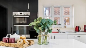 A modern kitchen with black cabinets, stainless steel appliances, and a white countertop. There are two vases with green plants, a tray with pastries, and a teapot. - Olympic House - Spacious house for a weekend with friends (Hinckley)