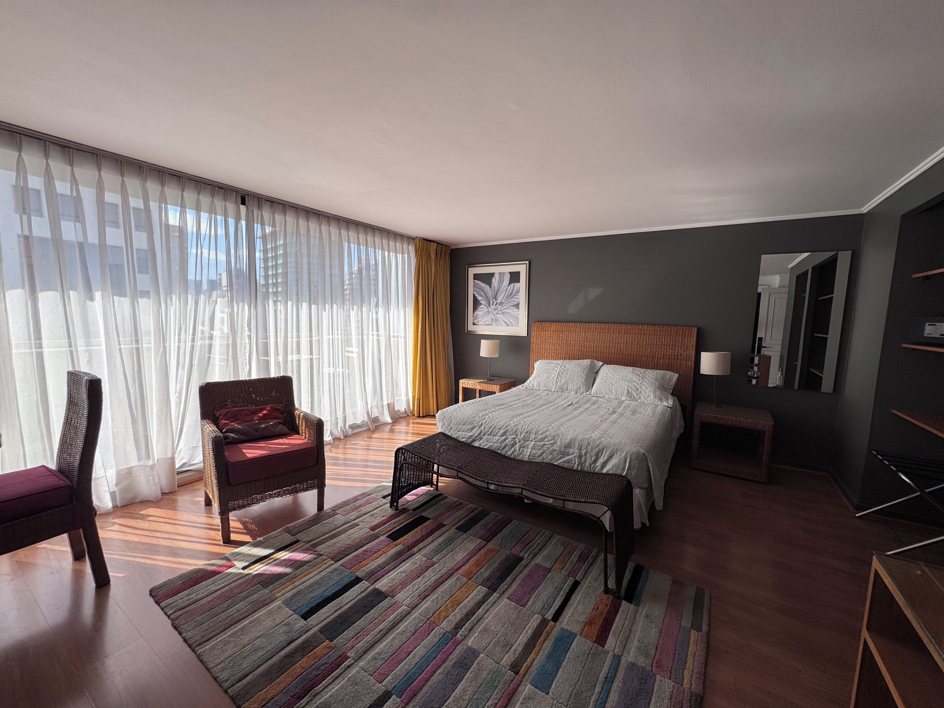 Una camera d'albergo con un letto grande, una sedia, una panca, un comodino, uno specchio e tende leggere.