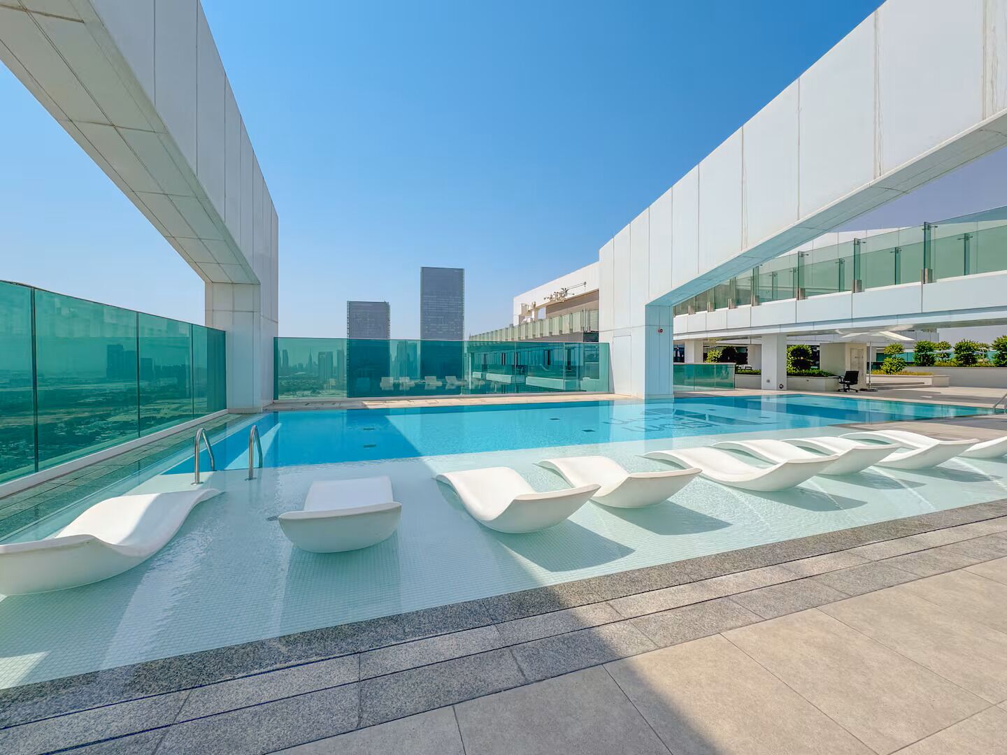 Ein Pool auf dem Dach mit gläsernen Wänden, modernen weißen Liegestühlen und Blick auf Wolkenkratzer.