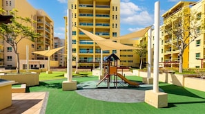 Parquinho com escorregador e estrutura de escalada, cercado por grama sintética verde e equipamentos de recreação bege, com prédios residenciais amarelos e de vários andares ao fundo.