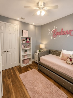 A bedroom with a bed, a nightstand, a dresser, a ceiling fan, and a wall with a name decoration.