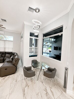 A modern living room with a sectional sofa, a glass dining table with chairs, a ceiling fan, and large windows.