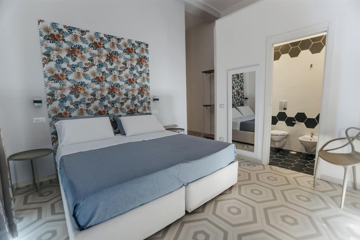 A modern bedroom with a bed, a nightstand, a chair, and a bathroom visible through an open door.