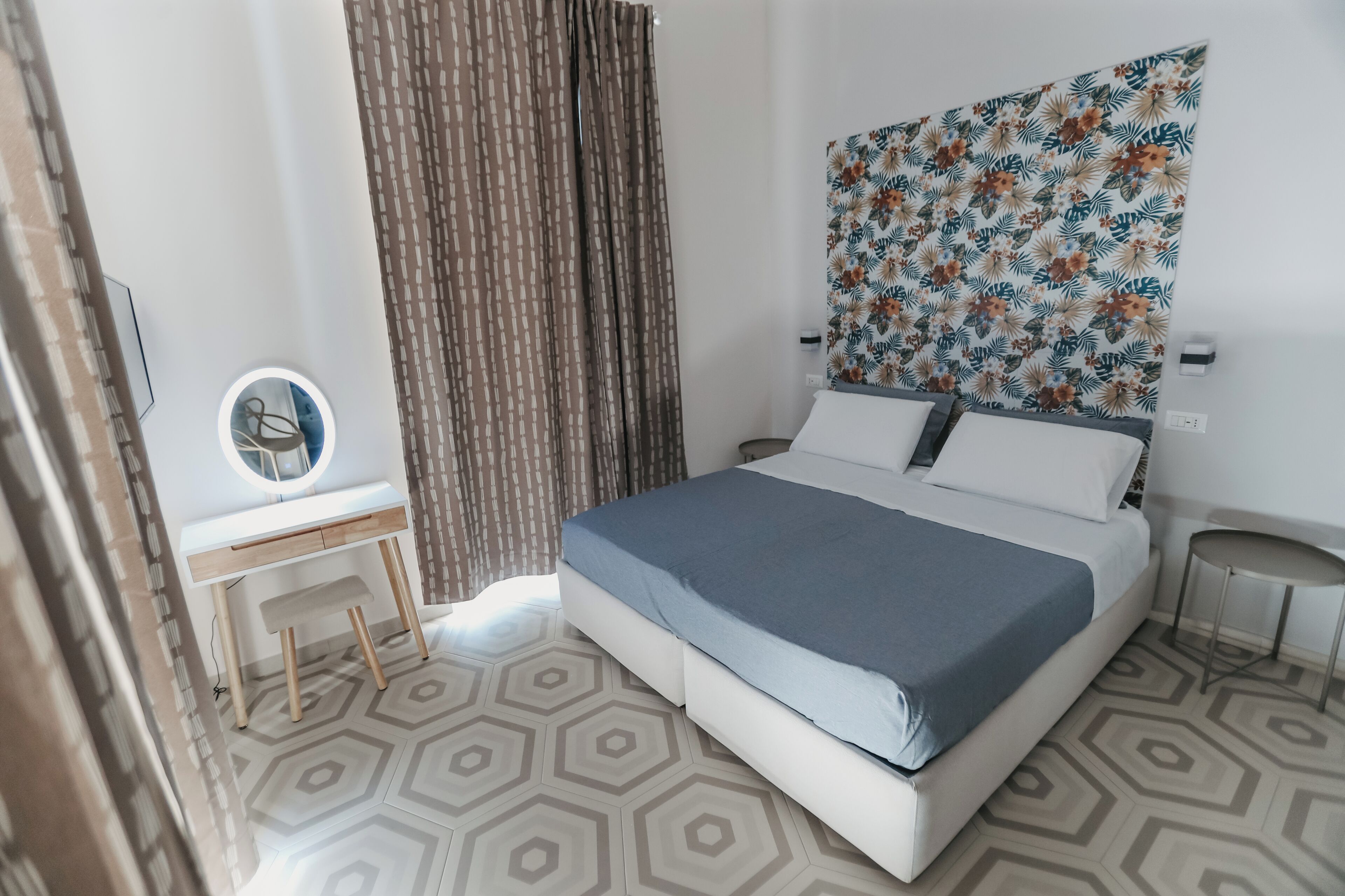 Una habitación de hotel moderna con una cama grande, un escritorio pequeño, un espejo redondo y un tapiz de pared con motivos.