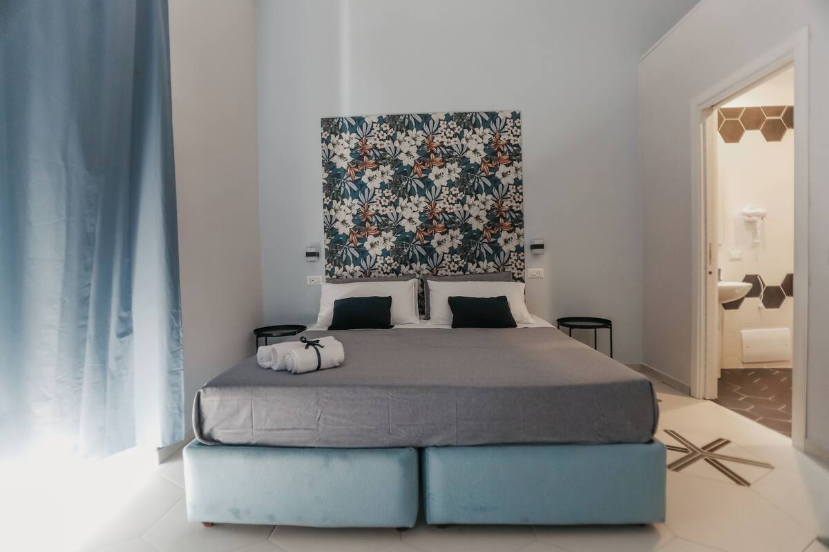 A modern bedroom with a large bed, a floral wall art piece, and a bathroom visible through an open door.
