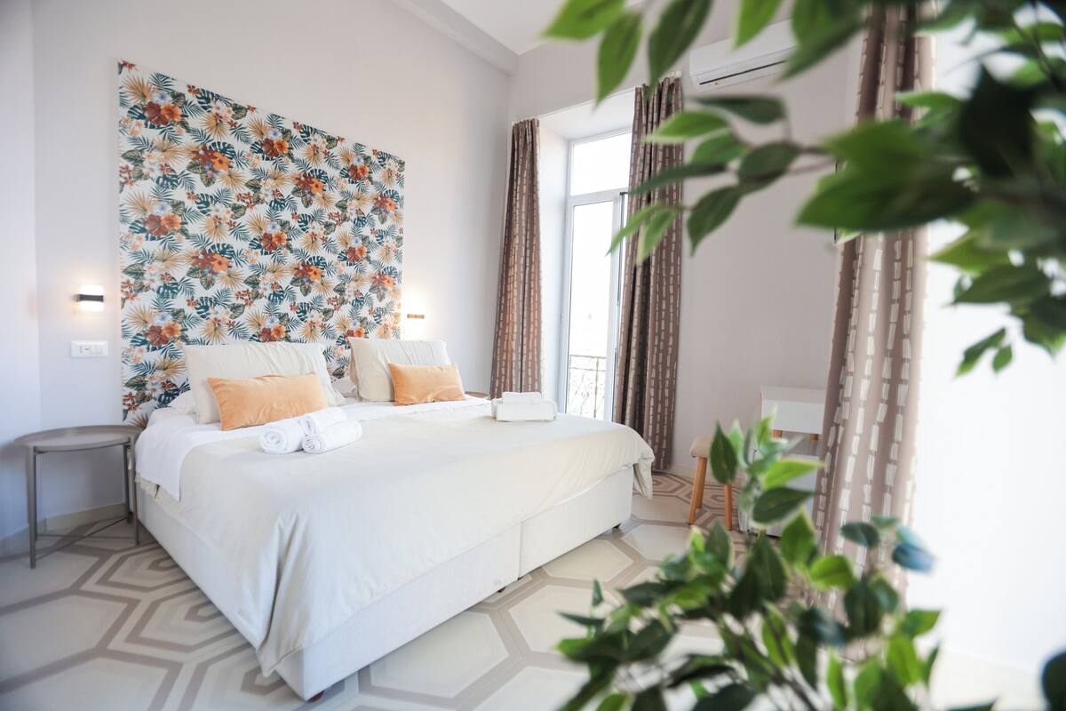 A modern hotel room with a large bed, a decorative wall tapestry, and a view of the outdoors.