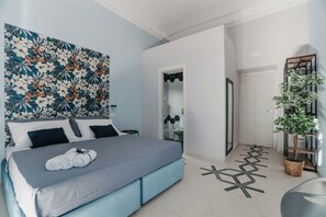 Ένα σύγχρονο δωμάτιο ξενοδοχείου με ένα μεγάλο κρεβάτι, μια κεφαλή κρεβατιού με σχέδια λουλουδιών, ένα φυτό σε γλάστρα και ένα γεωμετρικό σχέδιο στο δάπεδο.