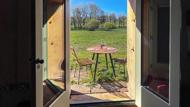 Eine geöffnete Tür, die auf eine Terrasse mit Tisch und Stühlen, eine Rasenfläche und Bäume im Hintergrund führt.