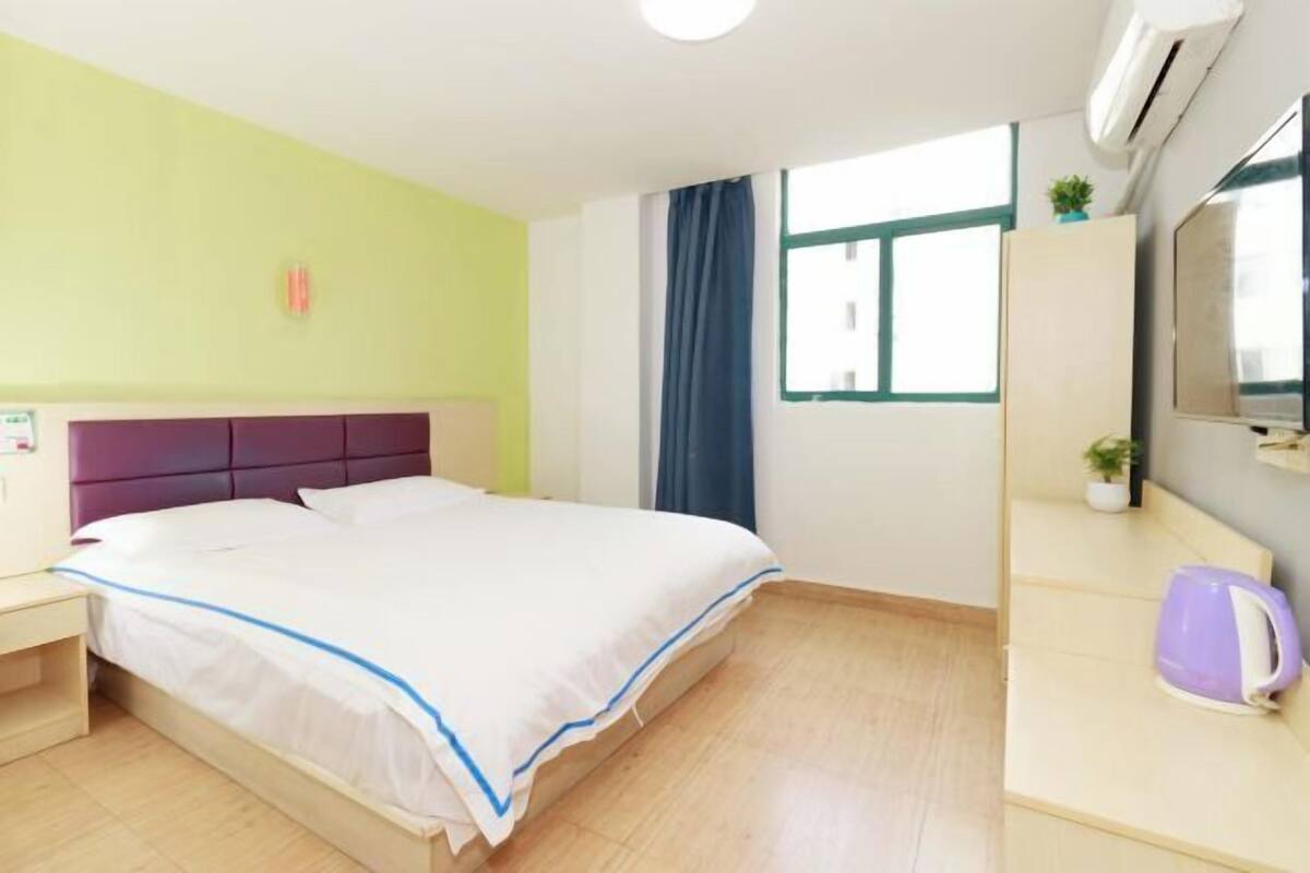 A hotel room with a bed, a bedside table, a small desk with a chair, a window with curtains, and a wall-mounted air conditioner.