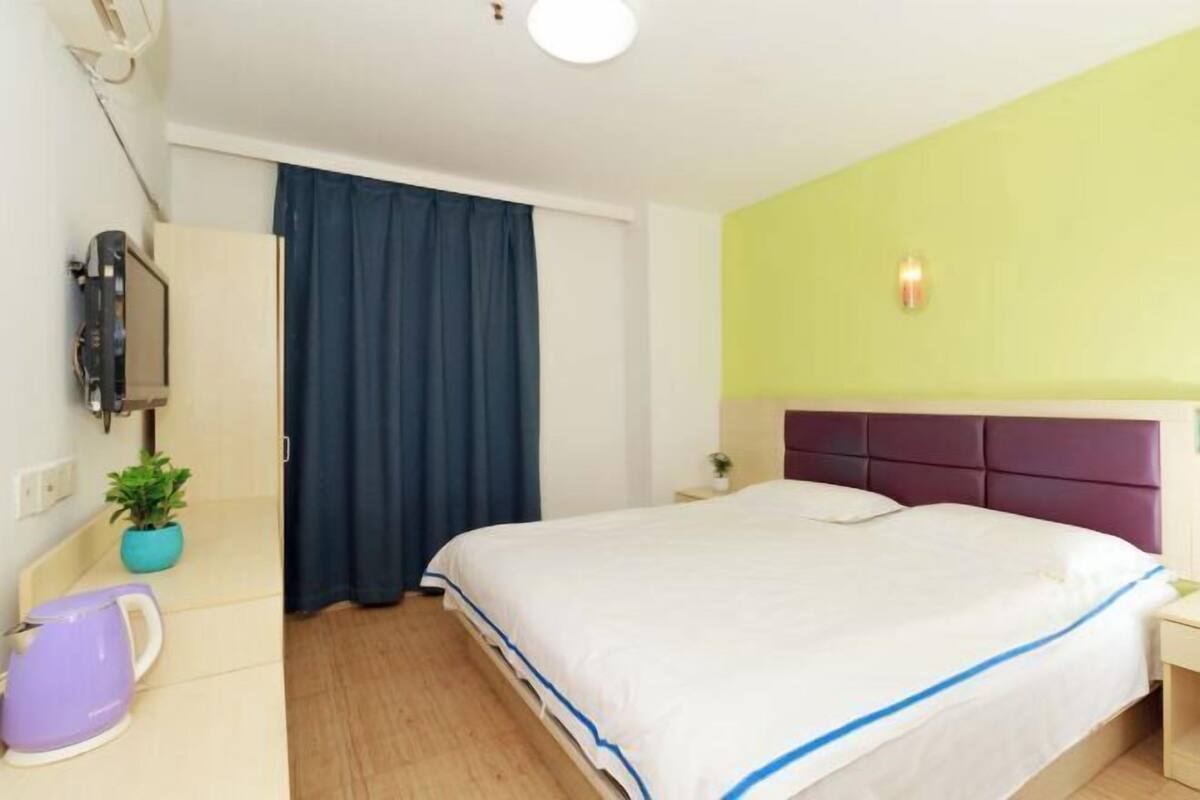 A hotel room with a bed, a nightstand, a television, a window with curtains, and a small plant.
