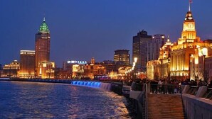 Un paisaje urbano frente al agua, con edificios iluminados, una destacada torre del reloj y un paseo marítimo de madera.