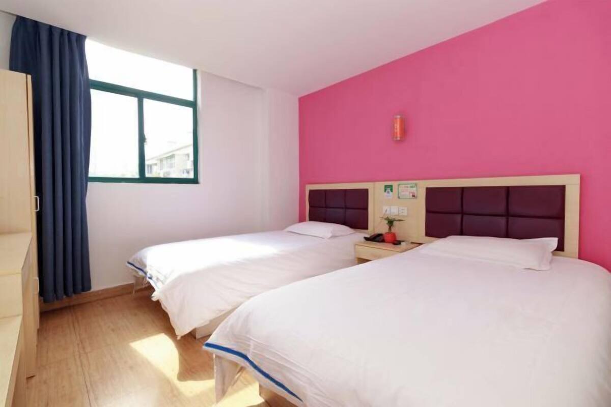 A room with two beds, a window with curtains, a pink wall, and a wooden floor.