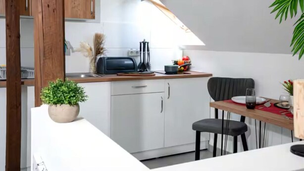 Een kleine keuken met witte kastjes, een houten aanrechtblad en een eethoek met een tafel en stoelen.