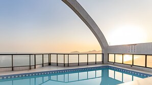 A swimming pool with a curved glass barrier, a clear view of the sea, and a sunset in the background. - ARK Houses - Flat Standard Vista-Mar c/ Piscina na Cobertura (Santos)
