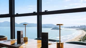 Habitación de hotel con un ventanal grande que ofrece vistas a una playa y montañas. Hay una mesa de madera con un cartel de número, una lámpara y un atril para el menú.