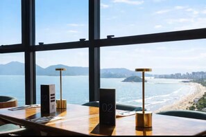 Hotelkamer met groot raam, uitzicht op strand en bergen. Er staat een houten tafel met een nummerbord, een lamp en een menuhouder.