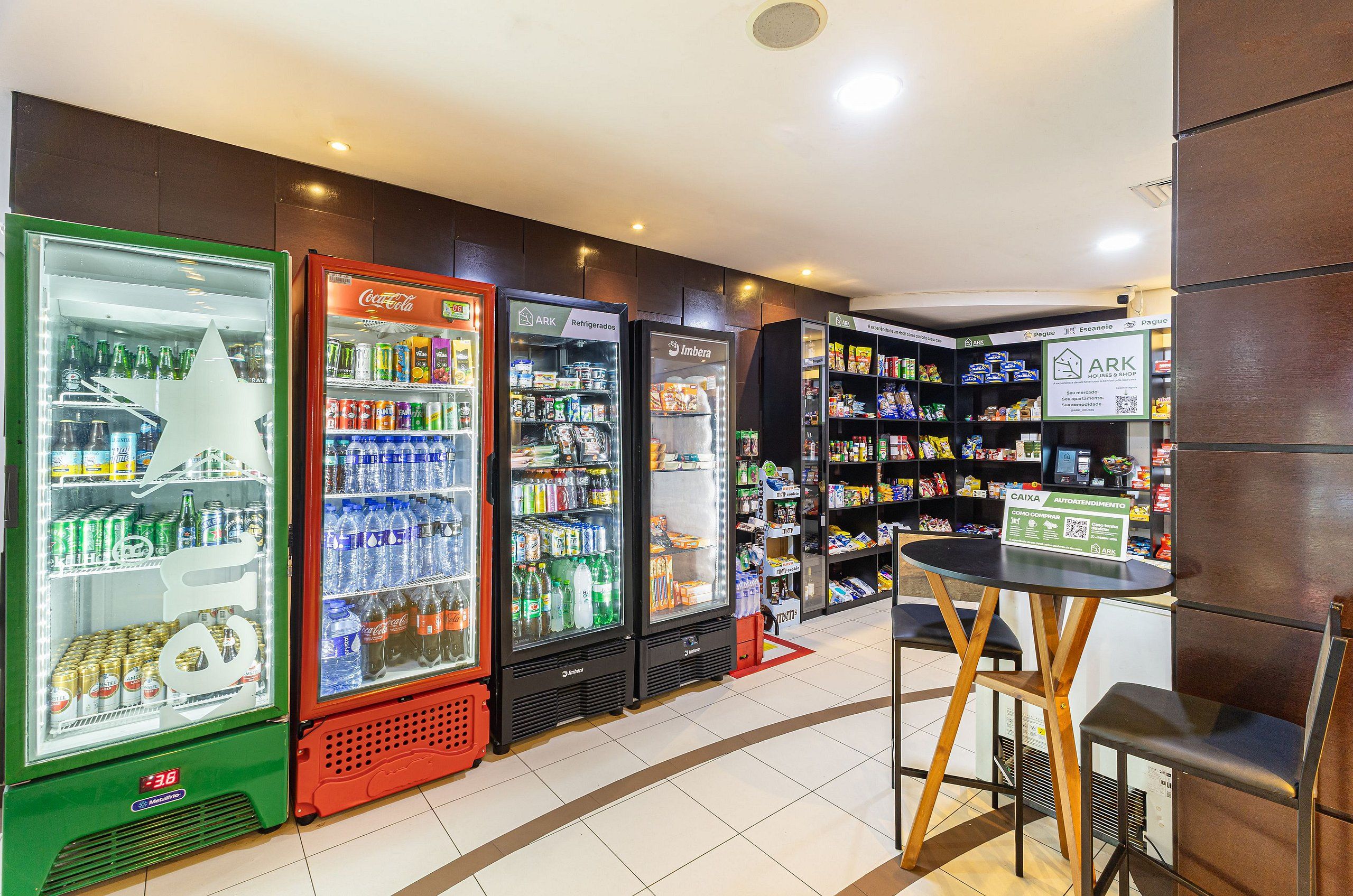 A convenience store with refrigerated beverage dispensers, shelves stocked with snacks and drinks, and a small seating area with a table and chairs.
